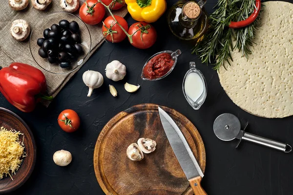 Tahta kesme tahtası ve geleneksel pizza hazırlama malzemeleri: peynir, domates sosu, mantar, zeytinyağı, biber, baharatlar. Siyah desenli masa arkaplanı. Üst Manzara. Durgun hayat. Boşluğu kopyala