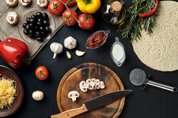 Tahta kesme tahtası ve geleneksel pizza hazırlama malzemeleri: peynir, domates sosu, mantar, zeytinyağı, biber, baharatlar. Siyah desenli masa arkaplanı. Üst Manzara. Durgun hayat. Boşluğu kopyala
