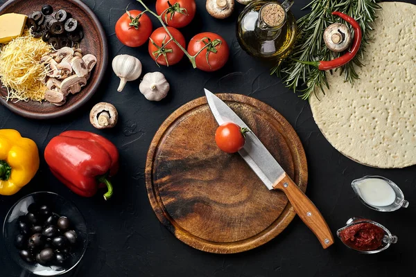 Tahta kesme tahtası ve geleneksel pizza hazırlama malzemeleri: peynir, domates, sos, zeytin, zeytin yağı, biber, baharatlar. Siyah desenli masa arkaplanı. Üst Manzara. Durgun hayat. Uzayı kopyala Düz