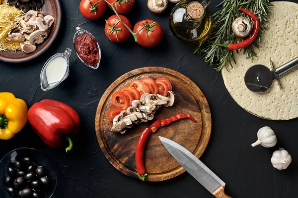 Tahta kesme tahtası ve geleneksel pizza hazırlama malzemeleri: peynir, domates, sos, mantar, zeytinyağı, biber, baharatlar. Siyah desenli masa arkaplanı. Üst Manzara. Durgun hayat. Boşluğu kopyala