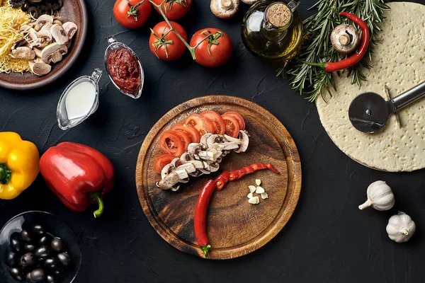 Tahta kesme tahtası ve geleneksel pizza hazırlama malzemeleri: peynir, domates, sos, mantar, zeytinyağı, biber, baharatlar. Siyah desenli masa arkaplanı. Üst Manzara. Durgun hayat. Boşluğu kopyala