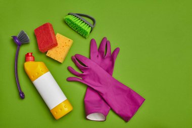 Yüksek kaliteli deterjanlar ve temizlik aksesuarları: sarı ve kırmızı süngerler, mor lastik eldivenler, boş etiketli dezenfektan ve yeşil arka planda iki modern fırça. Temizlik servisi