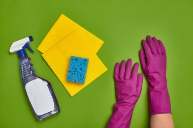 Profesyonel deterjanlar ve temizlik aksesuarları gibi: mavi sünger, boş etiketli sprey, mor lastik eldivenli eller ve yeşil arka planda sarı bulaşık bezleri. Temizlik hizmeti, küçük