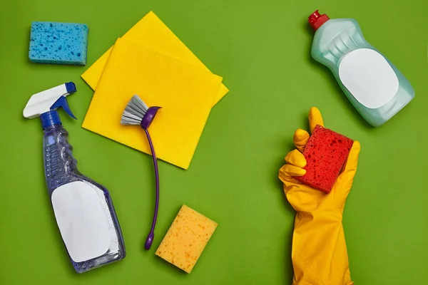 Modern detergents and cleaning accessories, such as: girl hand in a yellow rubber glove is holding a blue sponge. Cleanser for dishes with an empty label, yellow and blue sponge, spray, brush and