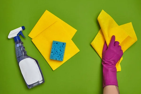 Modern detergents and cleaning accessories, such as: maiden hand in a purple rubber glove is holding a yellow dishcloth, spray with an empty label and blue sponge are laying nearby on a green
