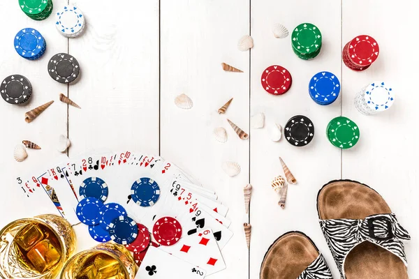 Card deck surrounded by poker chips and scattered seashells, glasses, flip flops, two glasses of whiskey with ice on white wooden background with copy space