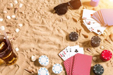 Plaj pokeri. Çipler ve kartlar kumsalda. Deniz kabuklarının, güneş gözlüklerinin ve soğuk bir bardağın etrafında. Üst Manzara. Uzayı kopyala Düz yatıyordu. Güneş parlaması