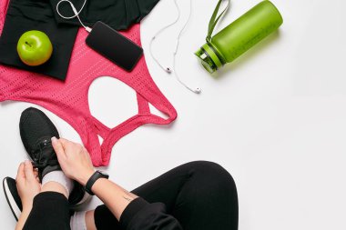 Spor taytlı genç kadın spor ayakkabıları giyiyor, eğitime hazırlanıyor. Spor aksesuarları, su şişesi, akıllı telefon, kulaklık, beyaz arka planda spor bilekliği düz görünüm yatıyordu.