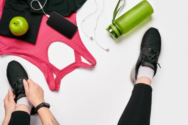 Spor taytlı genç kadın spor ayakkabıları giyiyor, eğitime hazırlanıyor. Spor aksesuarları, su şişesi, akıllı telefon, kulaklık, beyaz arka planda spor bilekliği düz görünüm yatıyordu.