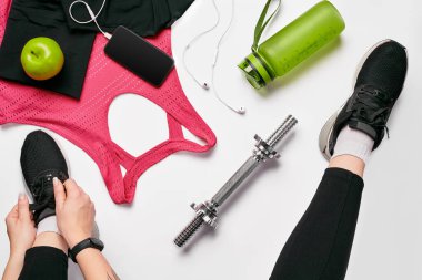 Young woman in sporting leggings laces sneakers, preparing for training. Accessories for sports, bottle of water, smartphone, headphones, fitness bracelet on white background flat lay top view