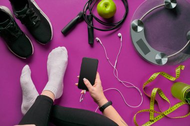 Young woman with mobile phone and accessories for sports, bottle of water, headphones, fitness bracelet, dumbbells on color background flat lay top view. Fitness concept