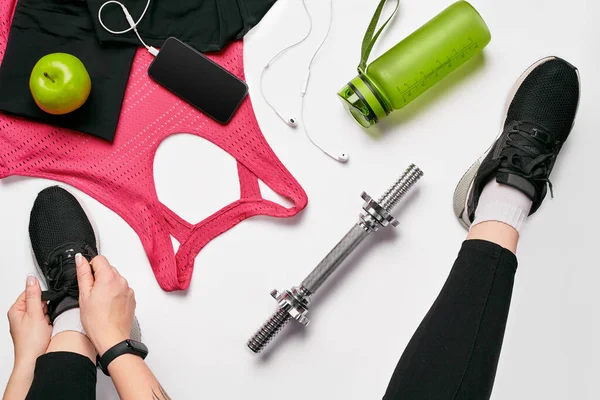 Spor taytlı genç kadın spor ayakkabıları giyiyor, eğitime hazırlanıyor. Spor aksesuarları, su şişesi, akıllı telefon, kulaklık, beyaz arka planda spor bilekliği düz görünüm yatıyordu.