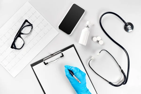 Doctor s table, tools, medical instruments, stethoscope, work in hospital on white background flat lay. The doctors hand in a blue glove records the indicators on a white paper blank. Top view. Still