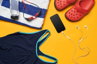 Mavi mayo, yüzme gözlüğü, çizgili havlu, kırmızı terlik, beyaz kulaklıklı siyah akıllı telefon ve sarı arka planda su şişesi gibi havuz aksesuarları. Renkli plaj