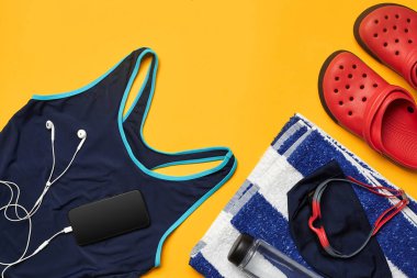 Mavi mayo, yüzme şapkası, gözlük, çizgili havlu, beyaz kulaklıklı siyah akıllı telefon, şeffaf şişe ve sarı arka planda kırmızı terlik gibi havuz aksesuarları düz yatıyordu.