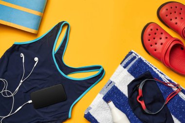 Mavi mayo, yüzme şapkası ve gözlükler, çizgili havlu, beyaz kulaklı siyah akıllı telefon ve sarı arka planda kırmızı terlik gibi havuz aksesuarlarının üst görünümü. Renkli plaj giyimi. Kopya