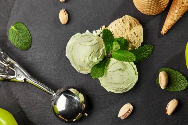 Nefis bir fıstıklı ve naneli, waffle külahlı ve saçılmış fındıklı çikolatalı dondurmanın yakın plan çekimi siyah bir arkaplan üzerinde servis edilir. Yakınlarda metal bir kepçe var.