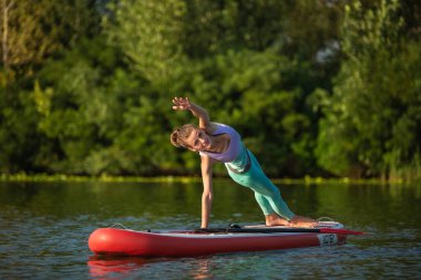Genç bir kadının ayakta duran bir panoda el ele durduğu fotoğraf. Üzerinde tayt ve bluz var. Güneşli bir gün, mavi bir göl ve sahilde yeşil ağaçlar.