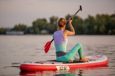 Sportif kadın üzerinde yoga yaparken paddleboard, yoga konumda kurulu sup, esneklik ve kas germe için egzersiz. Pratik yoga kürek gemide sabah kadın