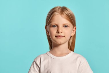 Close-up studio shot of a beautiful blonde little female with a long hair, in a white t-shirt, looking at the camera while posing over a blue background with copy space. Concept of a happy childhood.