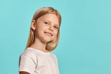 Close-up studio shot of a beautiful blonde little lady with a long hair, in a white t-shirt, looking at the camera and smiling while posing over a blue background with copy space. Concept of a happy
