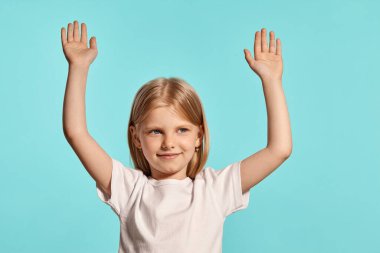 Close-up studio shot of a beautiful blonde child with a long hair, in a white t-shirt, raised her hands up and looking away posing over a blue background with copy space. Concept of a happy childhood.