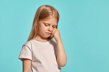 Close-up studio shot of a wonderful blonde little girl with a long hair, in a white t-shirt, crying posing over a blue background with copy space. Concept of a happy childhood.