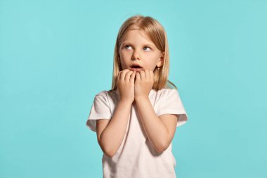 Close-up studio shot of a cute blonde kid with a long hair, in a white t-shirt, looking scared while posing over a blue background with copy space. Concept of a happy childhood.