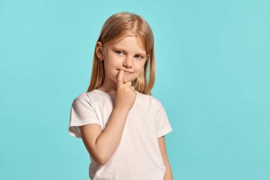 Close-up studio shot of a cute blonde little girl with a long hair, in a white t-shirt, looking shy while posing over a blue background with copy space. Concept of a happy childhood.