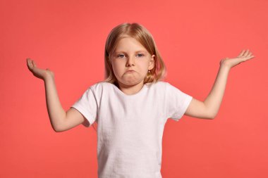 Close-up studio shot of a pretty blonde kid with a long hair, in a white t-shirt, acting like she does not know something while posing over a pink background with copy space. Concept of a joyful