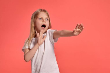 Close-up studio shot of a charming blonde little female with a long hair, in a white t-shirt, looking scared while posing over a pink background with copy space. Concept of a joyful childhood.