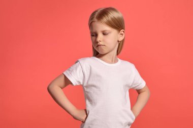 Close-up studio shot of a charming blonde little lady with a long hair, in a white t-shirt, looking disappointed while posing over a pink background with copy space. Concept of a joyful childhood.