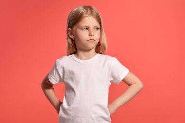 Close-up studio shot of a charming blonde kid with a long hair, in a white t-shirt, looking upset while posing over a pink background with copy space. Concept of a joyful childhood.