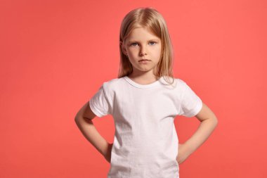 Close-up studio shot of a charming blonde child with a long hair, in a white t-shirt, looking unhappy while posing over a pink background with copy space. Concept of a joyful childhood.