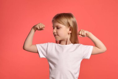 Close-up studio shot of a charming blonde little girl with a long hair, in a white t-shirt, showing her muscles while posing over a pink background with copy space. Concept of a joyful childhood.