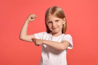 Close-up studio shot of a beautiful blonde child with a long hair, in a white t-shirt, showing her muscles and smiling while posing over a pink background with copy space. Concept of a joyful