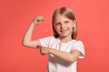Close-up studio shot of a beautiful blonde little girl with a long hair, in a white t-shirt, showing her muscles while posing over a pink background with copy space. Concept of a joyful childhood.