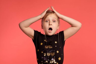 Close-up shot of a pretty blonde child in a combined dress, acting like she is shocked while posing against a pink background with copy space. Concept of a happy childhood.