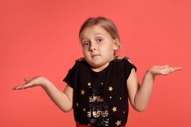 Close-up shot of a charming blonde little girl in a combined dress, acting like she does not know something while posing against a pink background with copy space. Concept of a happy childhood.