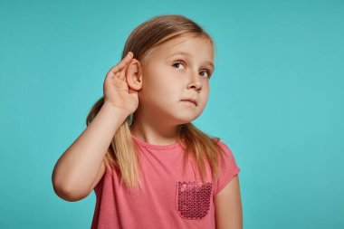 Close-up shot of a nice blonde little female with two ponytails on her head, in a pink dress, acting like listening to something while posing against a blue background with copy space. Concept of a