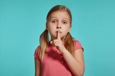 Close-up shot of a nice blonde child with two ponytails on her head, in a pink dress, showing sign quiet and looking at the camera while posing against a blue background with copy space. Concept of a