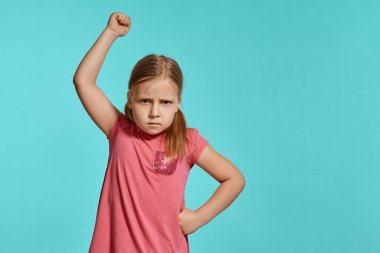 Close-up shot of a cute blonde little lady with two ponytails on her head, in a pink dress, being mad about something and raising her hand up while posing against a blue background with copy space