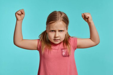 Close-up shot of a cute blonde kid with two ponytails on her head, in a pink dress, being unhappy about something and raising her hands up while posing against a blue background with copy space