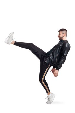 Full-length photo of a modern performer in glasses, black leather jacket, t-shirt, sports pants and light sneakers fooling around in studio. Indoor photo of a nice fellow dancing and raising his leg