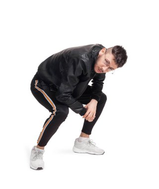 Full-length photo of a modern performer in glasses, black leather jacket, t-shirt, sports pants and light sneakers fooling around in studio. Indoor photo of a nice man dancing and looking at the