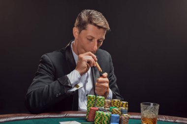 Arrogant male in a black slassic suit and white shirt is playing poker sitting at the table at casino in smoke, against a white spotlight. He rejoicing his victory smoking a cigar. Gambling addiction