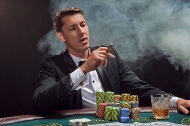 Arrogant man in a black slassic suit and white shirt is playing poker sitting at the table at casino in smoke, against a white spotlight. He rejoicing his victory smoking a cigar and looking down