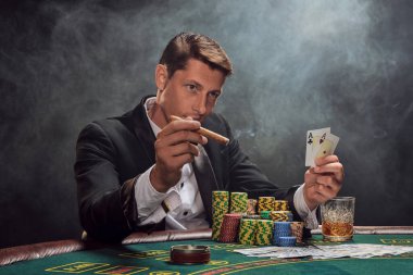 Smart male in a black slassic suit and white shirt is playing poker sitting at the table at casino in smoke, against a white spotlight. He rejoicing his victory showing two aces in his hand, smoking a