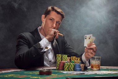 Smart man in a black slassic suit and white shirt is playing poker sitting at the table at casino in smoke, against a white spotlight. He rejoicing his victory showing two aces in his hand, smoking a