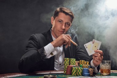 Elegant person in a black slassic suit and white shirt is playing poker sitting at the table at casino in smoke, against a white spotlight. He rejoicing his win showing two aces in his hand, smoking a